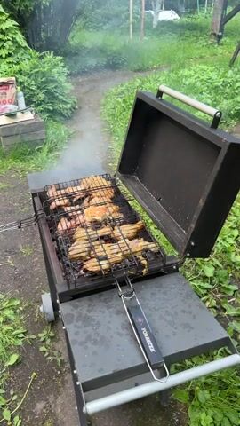 The video shows meat grilling on a Forester brand charcoal grill outside. The grill is black and gray with an open lid, revealing chicken and sausage pieces cooking over hot coals, emitting smoke. The grill sits on a gray concrete patio surrounded by green grass, plants, and a path. A wooden structure is partially visible in the background. A child's voice is heard in the background.