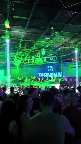 The video depicts a lively nightclub environment with a large crowd of people dancing and enjoying the music. The focal point is a DJ booth with a group of people behind it, possibly performers or DJs. Behind them, a large screen displays the word "TERMINAL" with a stylized "K" logo. Lighting is prominent, with bright blue and green lights illuminating the stage and crowd. The ceiling is visible, featuring a grid-like structure with various lights and fixtures. The overall atmosphere is energetic and festive.