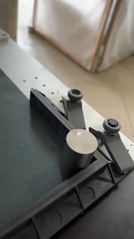 This close-up video shows a metal cylinder being laser-marked. The cylinder has a circular mark engraved on its top surface, which appears to be a logo or symbol. The cylinder is held in place by two dark grey, adjustable clamps, mounted on a dark green surface. A black 3D-printed fixture with circular cutouts is positioned in the foreground, likely to hold the cylinder or guide the laser. A metal ruler or track with visible holes runs along the top edge of the frame.

The activity is the laser engraving of the metal cylinder. The laser beam is visible as a bright point of light, actively etching the mark onto the cylinder. The process appears to be precise and controlled.

There are no people visible in the media file. The setting seems to be a workshop or manufacturing environment, given the tools and equipment present. The lighting suggests it is daytime, but no specific time of day can be determined. There are no overt emotional cues.

Visible text includes what appears to be "G6" engraved on the cylinder, alongside the circular logo. The surrounding context suggests this might be a marking for a specific grade or specification. The location context provided is Severo-Vostochnye Sady, Russia.