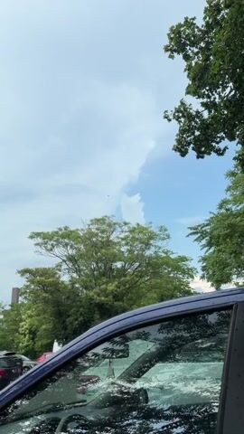 The video captures a partial view of a car's interior, reflecting a cloudy sky and trees. The sky is mostly overcast with patches of blue visible. Several trees with green foliage are present, some appearing in the foreground and others in the background. The car appears to be parked, as indicated by the presence of other vehicles in the distance. The front windshield and side window of the car are visible, with raindrops on the glass, suggesting it might be raining or has recently rained. The video is shot vertically, focusing on the sky and trees through the car's windows.