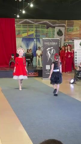 The video shows two young girls walking down a blue carpeted runway in what appears to be a fashion show. The girl on the left is wearing a red dress with white trim and a matching headdress. The girl on the right is wearing a black dress with a white sash. Behind them, there are several adults standing and watching. One person is playing an accordion. There is a banner in the background that says "MAMI MODELS". The overall environment looks like an indoor event space with lights and decorations.