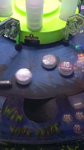 Here’s a description of the video:

The video shows a coin pusher arcade game. Several round tokens with the number “20” on them are being pushed around on a graveyard themed play surface. The game’s lower front panel reads “Win Your Prize” with indicators for 500, 100, and 20 tickets. A rotating cylinder of white light is visible in the background, and the game appears to be in motion.