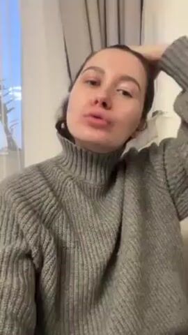A woman with dark hair and light skin is standing inside. She is wearing a gray turtleneck sweater. She pulls her hair back from her face and holds it in her hands. In the background, a window with white curtains is partially visible.