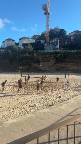 A group of people are playing beach volleyball on a sunny day. There are about ten individuals in the sand court, appearing to be engaged in a competitive match. The volleyball net is clearly visible, with orange lines marking the boundaries of the court. A construction crane towers in the background, adding a modern element to the beach scene. Behind the beach, houses are built on a small cliff, and foliage is growing. The sky is a bright blue with scattered clouds. The camera angle is from an elevated position, looking down onto the beach. A railing partially obstructs the bottom of the frame.