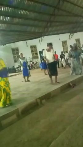 In a wide shot taken from slightly below eye level, at least nine adults and one child can be seen gathered on a gray concrete floor in a large, indoor space with a corrugated metal roof. The majority of the adults are women, many wearing head coverings and colorful patterned dresses or skirts. One woman stands out in a bright yellow and blue floral print dress. A man in a red shirt is seen near the back of the space.

The group is dancing to music. The people move in a relaxed, informal way, seemingly enjoying the music and each other's company. The large indoor space appears to be some type of assembly hall or event space, with white walls and windows visible along one side. The concrete floor is slightly raised, with a visible step in the foreground. The overall lighting is somewhat dim, but the space is still well-lit enough to clearly see the people and their movements. The child in the background appears to be watching the adults.