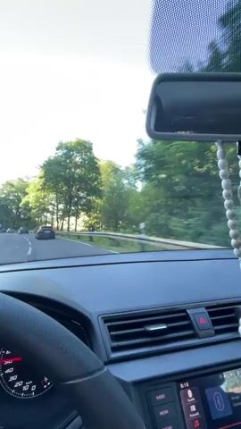 A first-person perspective video captured from inside a car. The driver's hand is visible on the steering wheel, and the dashboard is in view, showing a speedometer reading around 51-52 km/h. The car is on a road surrounded by trees and greenery, with other cars visible ahead. The sky is clear and blue, suggesting daylight. The camera pans slightly, offering a view of the passing scenery.