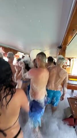 The video captures a lively foam party taking place on the deck of a boat, likely off the coast of Menderes, Turkey. A diverse group of young to middle-aged adults, both men and women, are immersed in a thick layer of white foam that covers the boat's floor and their bodies.

Most of the men are shirtless or wearing swim trunks, while the women are dressed in bikinis or swimwear. The atmosphere is jubilant and energetic, with individuals dancing, clapping, and smiling broadly. A central figure, a woman in a blue and white striped bikini, stands out with her infectious smile and enthusiastic gestures, raising her arms in enjoyment. In closer shots, a blonde woman wearing sunglasses and a beige net-like cover-up smiles directly at the camera, accompanied by a man next to her.

The boat's interior appears to be a covered deck area, featuring wooden structures and railings. Bright sunlight streams in, suggesting it is daytime, and glimpses of clear turquoise water can be seen through open sections of the boat, indicating its coastal setting. A large, glowing white inflatable object or light fixture is visible amidst the foam in some shots, adding to the party ambiance. Everyone appears to be having a fun, celebratory time. No visible text is present in the video.