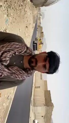 A vertical video shows a man in the foreground, wearing a dark green long-sleeved shirt and a light-colored plaid scarf wrapped around his neck. He has dark hair and a beard, and he is looking directly at the camera, speaking.

Behind him, a newly paved asphalt road stretches into the distance. To the right of the road, there are rough, dusty patches of ground and some low-rise buildings, some of which appear to be under construction or in disrepair. In the mid-ground on the road, heavy construction machinery, possibly a road roller or paver, is visible, indicating ongoing road work. Further down the road, another vehicle, possibly a white car, is visible. The sky is bright and clear, suggesting a sunny day. Mountains can be seen in the far distance. The overall scene depicts a road construction project in an arid, possibly rural, environment. The man is likely addressing the condition of the road or the ongoing work. The setting is Kandahar, Afghanistan.