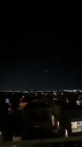 The video shows a night cityscape with a dark sky. In the distance, city lights are visible on the horizon. Closer in, the outlines of houses and other buildings can be seen. Above the cityscape, a distant light is seen in the sky. As the video zooms in on the light, it is revealed to have three distinct points: one green, one white, and one red. After a moment, the video zooms back out to the general cityscape.