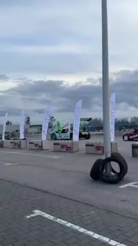 The video shows a paved outdoor course with several flags lined up along the edge, each bearing the name "PETER." Behind the flags are concrete barriers topped with orange cones. A green and white racing truck is visible beyond the barriers, with a person walking near it. To the right of the flags, a stack of tires is present, and a red car is parked further down the course. The sky above is overcast with a mix of grey and white clouds. The camera is static, providing a fixed perspective of the scene.