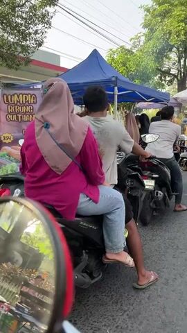 The video depicts a street scene with several motorcycles and people. A blue awning tent is set up, with people interacting around it. A woman in a pink shirt and hijab is visible behind the tent, appearing to be serving someone. Another woman in a brown hijab is also present. A man wearing a light-colored shirt and dark pants is sitting on a black motorcycle. In the foreground, another motorcycle is partially visible. Further down the street, a red motorcycle is parked, and people are standing or sitting under umbrellas. Several motorcycles are parked along the street. The environment appears to be outdoors, possibly a market or roadside stall. The overall impression is of a busy street scene with vendors and customers.
