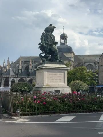 This is a video of a statue of Abraham Duquesne in a public square. The statue is made of bronze and depicts Duquesne in military attire, holding a sword and a pair of binoculars. He is standing on a pedestal with an inscription on it. In the background, there is a large building that appears to be a church or cathedral, with gothic architecture. There are also some trees and rose bushes in front of the statue. The sky is cloudy.