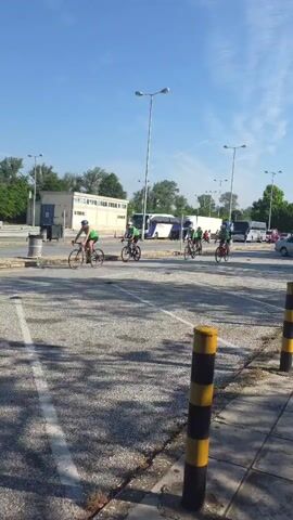 A group of five cyclists on road bikes wearing green shirts and helmets rides left to right across a paved area with a few parked buses in the background, one cyclist turning to ride away from the camera. The cyclists are riding on a bright sunny day. The camera pans with the cyclists as they ride.