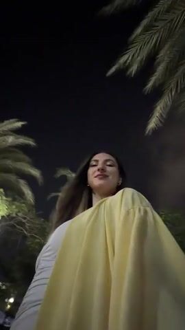 A woman walks at night outdoors, wearing a white strapless top and a yellow shawl. Palm trees are visible in the background, along with streetlights and other people walking in the distance. The camera is pointed upwards towards the woman as she walks, then pans to a selfie-style shot where she smiles and looks at the camera.