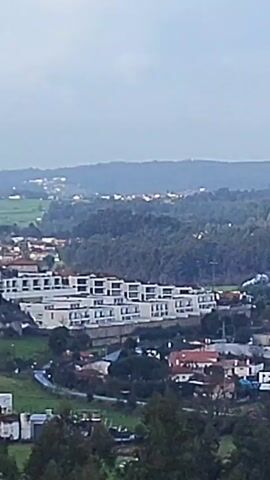 The video shows a panoramic view of a rural area with a mix of residential buildings, green fields, and dense forests. The buildings vary from modern, multi-story apartments to smaller, traditional houses with red tile roofs. The landscape slopes upward into a series of hills, and the sky is overcast with a muted, grey tone. The camera slowly pans from left to right, revealing more of the sprawling landscape, including some electrical pylons in the fields.