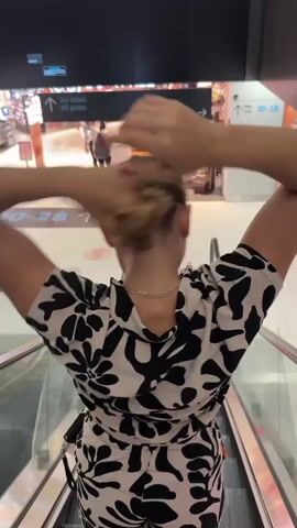 A woman stands on an escalator, facing away from the camera. She has blonde hair and is wearing a black and white patterned shirt. She is in the process of tying her hair up into a bun, using her hands to gather and twist it. In the background, there are signs indicating "All gates" and various shops. The escalator is moving downwards, as evidenced by the motion blur in the background.