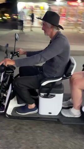 Two people are riding on a three-wheeled electric scooter on a street at dusk. The person in the front appears to be an older male wearing a hat and sunglasses. The person in the back is wearing a black hooded sweatshirt. The scooter has a large sign attached to the back that advertises a show and disco. Several cars are parked and driving on the street. Trees line the side of the street.