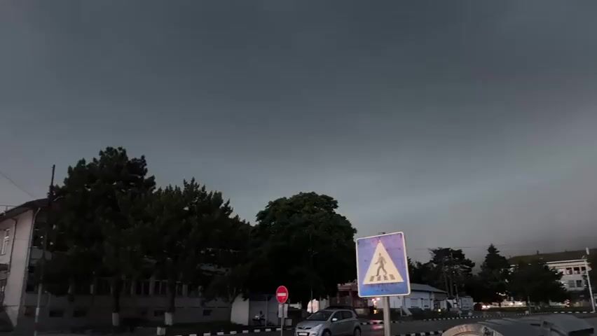 A dark, ominous sky with visible clouds dominates the upper half of the frame. In the foreground, a blue and white pedestrian crossing sign is prominent on the right. Below the horizon, a street with light traffic, including at least two cars, is visible. On the left side, two multi-story buildings stand, partially obscured by several large, dark green trees. The overall scene suggests an impending storm or a heavily overcast day.