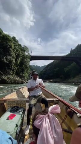 The video shows a man rowing a boat down a river. He is wearing a white t-shirt and shorts. The boat is filled with people, and they are all looking at the camera. The river is surrounded by trees, and there is a bridge in the background. The sky is cloudy.
