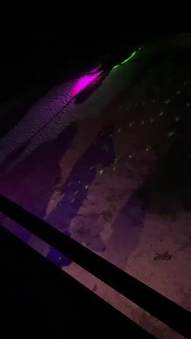 The scene is dark, illuminated by purple and green lights. The purple light shines on the ground, creating long, distorted shadows of at least three people. A black railing is visible in the foreground. Loud music plays in the background.