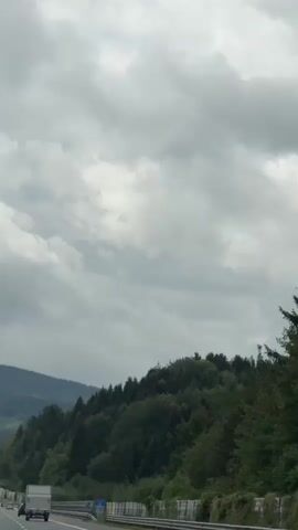 A car is driving on a highway. The sky is cloudy and gray. There are trees and hills in the background. There are other cars on the highway, including a truck and a black car. The road is paved and there are white lines marking the lanes. There are barriers on the side of the road.
