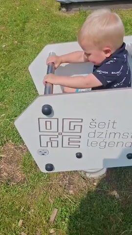A blonde toddler wearing a dark, hooded shirt with white lettering sits inside a playground ride with metal handles. The ride is shaped like an arrow with a white panel on top displaying text and a graphic. The text reads "ŠEIT DZIMST LEGENDA" with the word "OGRE" in large, stylized letters to the left of the text. The ride is situated on a grassy area with some fallen leaves and sticks. The toddler is holding onto the metal handles and appears to be looking down. The video appears to be a short clip from a smartphone, likely a vertical recording.