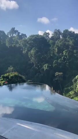 The video begins with a close-up shot of an infinity pool overlooking a lush green jungle. A bird flies in the distance. The camera then pans to reveal a wooden deck with a railing, a sign, and stairs leading down. Further into the video, the camera shows an outdoor seating area with tables and chairs, some under a thatched-roof structure, and palm trees. Finally, the video moves to a more enclosed area with more tables and chairs, and a bar in the background. The overall atmosphere appears to be a resort or a high-end restaurant in a tropical setting. There are no people clearly visible in the early part of the video, but a few individuals can be seen in the background of the restaurant area, seemingly staff members.