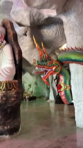 The video displays a dimly lit interior, possibly a cave or a temple, with rough, textured walls. In the foreground, a large statue of an elderly man with a long white beard and white robes sits on a rock formation adorned with gold coins. He holds gold coins in his lap and wears elaborate jewelry. To the right of this statue, a colorful, serpent-like statue with a dragon's head, adorned with ornate fins and scales, is prominently featured, appearing to coil around a rock pillar. In the background, other statues and figures are visible, including what appears to be a golden mask on a stand and other adorned figures. The lighting is dramatic, with pink and yellow spotlights illuminating sections of the scene, creating an ethereal atmosphere. The floor is reflective, suggesting it may be wet or polished stone. The camera movement is fluid, panning and tilting to capture different aspects of the elaborate display, creating a sense of exploration within the space.