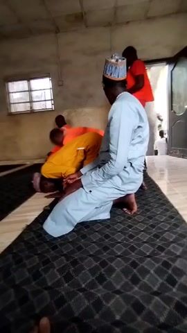Here is a concise description of the video:

The video shows a room with several men engaged in prayer. Three men are visible in the foreground, kneeling on a patterned rug. The man in the center is wearing a light blue robe and a matching hat, while the other two are wearing an orange jacket and a yellow shirt respectively. A window with a barred frame is visible on the left side of the frame. Another person wearing a red shirt is visible in the background. The room has light-colored walls, a tiled floor, and an open doorway.