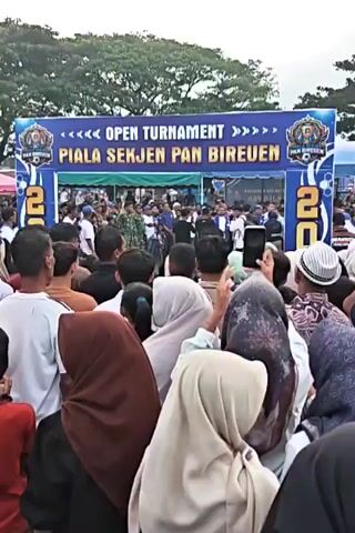 A group of people are gathered outdoors in front of a large blue and yellow banner that reads "OPEN TOURNAMENT PIALA SEKJEN PAN BIREUEN 20." The crowd is dense, with many individuals wearing hijabs. Some people are holding up cell phones, likely to take pictures or record video. Behind the banner, a group of people are visible, some wearing blue shirts and others in military-style uniforms. The overall scene suggests an event or competition is about to begin or has just concluded. The ground appears to be paved or a dirt area, and the background suggests an open outdoor space with some trees and structures in the distance.