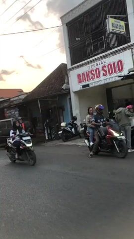 The video shows a street scene in what appears to be a market or a commercial area. On the right side of the frame is a building with a sign that reads "SPESIAL BAKSO SO." Several motorcycles are parked in front of this building. In the center of the frame, a person wearing an orange vest and a mask is walking. To the left of this person is a dark-colored van. The street is paved, and there are other buildings and structures visible in the background. The overall lighting suggests it might be late afternoon or early evening. A person on a motorcycle, partially blurred due to motion, is in the foreground on the right, moving away from the camera.