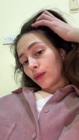 A young woman is shown in a close-up selfie video. She is light-skinned with dark brown hair that is slightly wavy. She is wearing a white undershirt and a mauve corduroy button-down shirt. The wall behind her is a light yellow color and has a light switch. She runs her hand through her hair and then states "I can't get up".