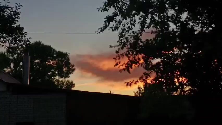 This video captures a sunset with colorful clouds. Trees and power lines are visible in the foreground, obscuring some of the view. A house with a dark roof can be seen in the distance. The camera appears to be moving, possibly from a vehicle.