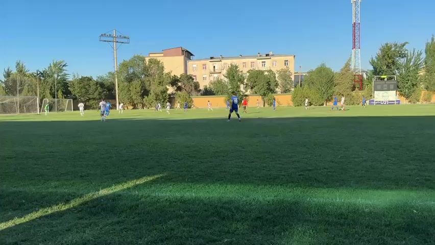 The video shows a wide shot of a soccer field during daylight. The sky is clear and blue, suggesting a sunny day. The field is covered in green grass, with a white line marking the edge of the field visible in the foreground. Several soccer players, wearing blue and white uniforms, are scattered across the field, engaged in a game. One player in a blue uniform stands out in the middle of the field, appearing to be a referee or a player in a central position. In the background, there are buildings, trees, and a tall communication tower. The players are in motion, dribbling the ball, running, and interacting with each other, indicating an active soccer match is in progress. The overall scene depicts a typical outdoor sporting event.