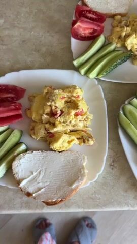 The video shows a top-down view of a breakfast spread on a speckled countertop. There are two white plates and one brown and white patterned plate, all containing food. The white plates hold scrambled eggs with visible red pepper pieces, slices of cucumber, and slices of tomato. One white plate also has a slice of bread spread with butter or cream cheese. The brown and white plate contains scrambled eggs, cucumber slices, tomato slices, and a slice of bread spread with butter or cream cheese. A portion of a microwave oven is visible in the background. The bottom edge of the frame shows the feet of a person wearing slippers.