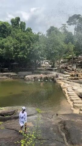 A person wearing a white shirt and a white cap stands on a rock at the edge of a pool of water. They are facing away from the camera with their arms raised, appearing to splash water. The water in the pool is greenish, and there are rocks and vegetation surrounding it. In the background, trees and a cloudy sky are visible, as well as a small waterfall and structures on the far side of the pool. The person's movement is captured in a short video loop.