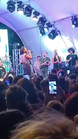A group of musicians perform on a stage under a tent. A female singer in the foreground is wearing a light-colored top and a dark skirt, holding a microphone and singing. To her right, a male musician plays a guitar. Behind them, another female singer, wearing a dark outfit, sings into a microphone, and a male musician with curly hair sings into a microphone. Several audience members are visible in the foreground, many holding up smartphones to record the performance. The stage is illuminated by bright stage lights.