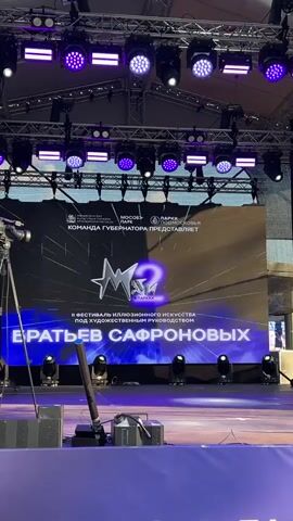 The video shows a large stage setup for an outdoor event. A large LED screen is prominently displayed, featuring the logo "M2" and text in Russian, indicating a festival of illusionary art. The screen also displays QR codes and sponsor logos. Above the screen, a complex lighting rig with purple LED lights is visible. To the left of the screen, a professional video camera on a tripod is positioned. Stage equipment, including speakers and lighting fixtures, are arranged on the stage floor. The background suggests an outdoor setting with a blue sky. The footage pans and zooms slightly, capturing different angles of the stage and screen.