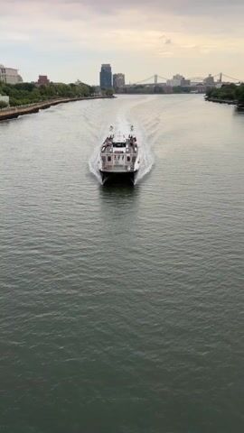 The video shows a catamaran ferry moving away from the camera on a body of water. The ferry has a white superstructure with two levels and is carrying passengers on the upper deck. People are visible standing and sitting on the deck, some appear to be talking to each other. The ferry is leaving a white wake behind it. In the background, a city skyline is visible with buildings and a bridge. The sky is overcast.