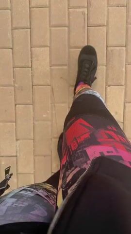 The media file shows a first-person perspective of someone walking on a paved sidewalk. The subjects are the person's legs and feet, clad in patterned leggings with a cityscape design and black sneakers. The scene is an outdoor setting with a clear view of the ground, composed of rectangular beige paving stones. The activity is walking.

Notable details include the vibrant, artistic pattern on the leggings, which appears to depict buildings with colorful lighting. The time of day seems to be daytime, given the lighting conditions, and the weather appears clear. The location cues, such as the paving stones and the general outdoor environment, suggest an urban or suburban area, possibly a park or a pedestrian walkway. There are no visible emotions conveyed as the focus is on the lower body and the ground. No visible text is present in the media file.