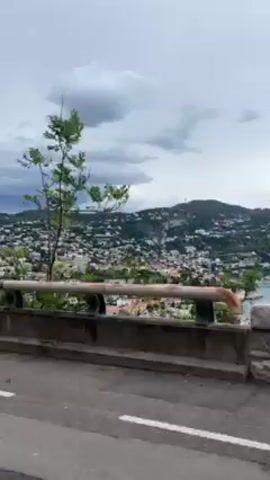 Here is a description of the video:

The video shows a view from a road or bridge with a concrete barrier. In the background, there's a densely populated hillside leading down to the water, likely a coastal town or city. The sky is overcast with dramatic cloud formations. Trees are visible in the foreground, partially obscuring the view. The road is asphalt with a white line marking the lane. The overall impression is of a scenic overlook on a cloudy day.