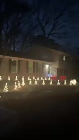 The video shows a two-story house at night, decorated with Christmas lights. The house is a mix of brick and siding, with dark shutters on the windows.  A line of small, lit-up Christmas tree decorations extends across the front lawn. There are also some red light decorations around the front porch area.  The sky is dark, and there are bare trees in the background, suggesting it might be late fall or winter.  The scene is dimly lit by the house lights and Christmas decorations.