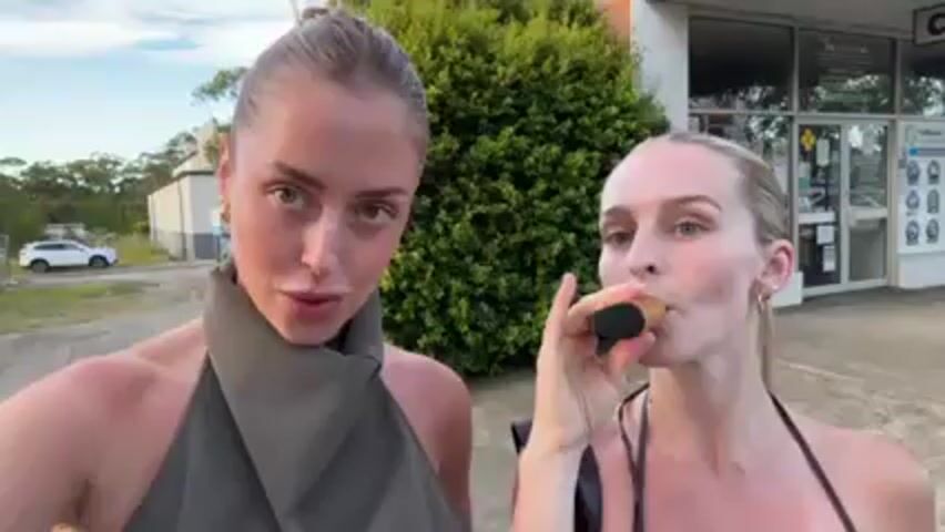 Two fair-skinned women are standing outside, looking at the camera. The woman on the left has her hair pulled back in a bun, is wearing a sleeveless green halter top, and makes a kissing face. The woman on the right is wearing a black top and has her hair pulled back into a ponytail. She is inhaling from a gold-colored vape pen. In the background are a bush, a white car, and a building with a sign on the window.