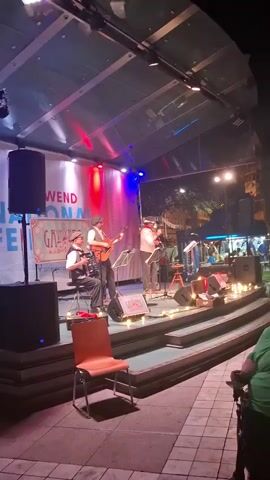 The video shows a musical performance taking place outdoors on a stage at night. The stage is set up with a backdrop banner that has text on it, though it is partially obscured. There are three musicians visible on stage. On the left, a person is seated and playing an accordion. In the center, a person is standing and playing an acoustic guitar. To the right of the guitarist, another person is standing and appears to be singing into a microphone, also holding a clarinet. All three musicians are wearing similar attire, including hats and vests, giving them a vintage or themed appearance. The stage is lit with red and blue lights, and there are string lights along the edge of the stage. In front of the stage, there are tiered seating areas or steps, with a chair visible in the foreground. The background shows trees and buildings, suggesting an urban or town setting. The overall atmosphere appears to be festive, likely a concert or festival.
