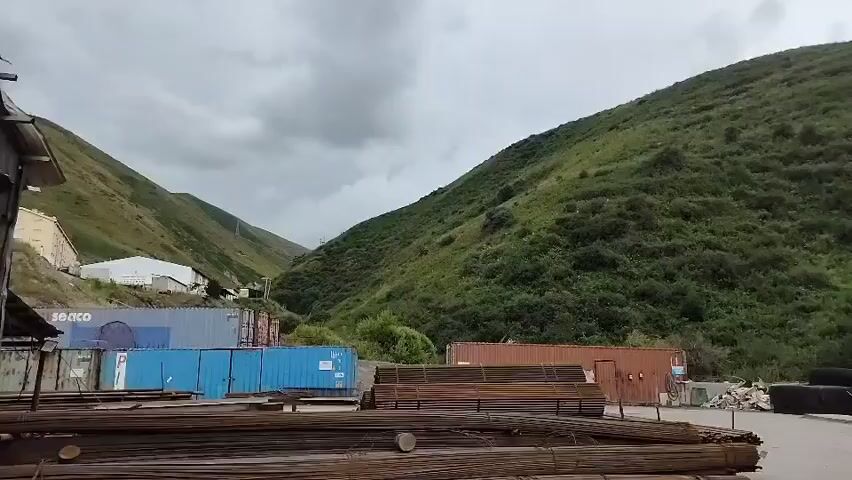 The video displays an outdoor industrial area situated in a valley with lush green hillsides under a cloudy sky. In the foreground, there are stacks of metal rebar and various shipping containers, some blue and one orange. Further back, a white building is visible on the left, nestled against the hillside. Two tall, slender trees stand prominently in the mid-ground. The overall impression is one of a functional, albeit somewhat rugged, operational site amidst a natural landscape.