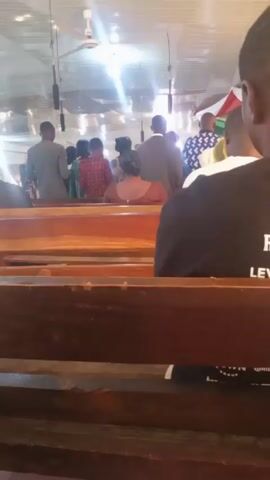 The camera is positioned from a low angle behind wooden church pews. In the background, a group of people stand facing away from the camera, likely towards the altar or front of the church. The individuals' clothing styles vary, including suits, dresses, and patterned shirts. Ceiling fans and hanging microphones are visible above the crowd, along with a brightly lit white ceiling that reflects the sunlight. The person filming is sitting in the pew and wearing a black t-shirt with white lettering.