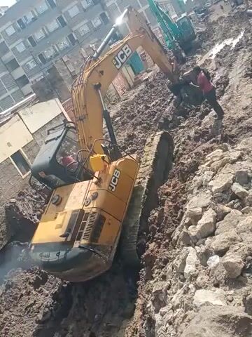 The video displays an active excavation site, likely in Mugumo-ini ward, Kenya. A large yellow JCB excavator is prominently positioned within a deep trench, which has uneven, rocky, and muddy sides. The excavator's arm is partially extended, indicating ongoing earth-moving operations. Another green excavator can be seen in the background, further along the trench. A person wearing a pink shirt is visible standing inside the trench, near the yellow excavator, possibly supervising or assisting with manual labor. The scene is brightly lit, suggesting it is daytime, with clear skies. Residential or commercial buildings are visible in the distant background, placing the construction within an urban or peri-urban environment. The visible text "JCB" clearly identifies the brand of the main excavator.