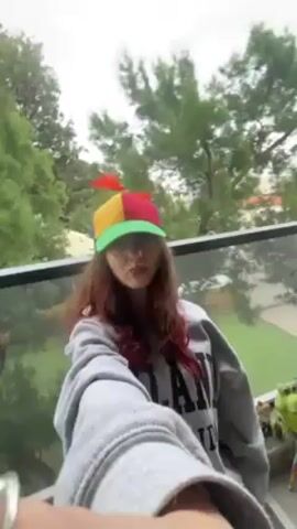 A person, possibly a young woman with reddish-brown hair, is taking a selfie-style video on what appears to be a balcony or porch. They are wearing a grey sweatshirt with black lettering and a multicolored propeller hat. The propeller on the hat is spinning rapidly. Behind the person is a glass railing, green trees, and a building in the distance. The sky is overcast.  There are decorative objects on the ground to the right.