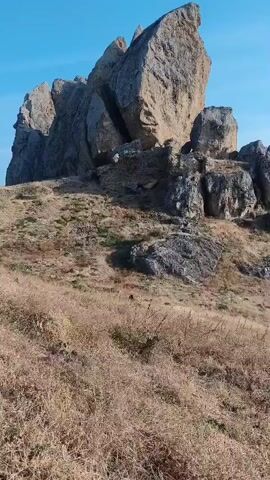 A video shows a view of rocky hills and a body of water under a clear blue sky. The sun is visible, creating a bright glare on the water. The landscape appears dry and arid, with sparse vegetation. In the foreground, tall, dry grass dominates the scene. In the background, several large rock formations are visible, with one particularly prominent, jagged peak. The video pans across the landscape, offering a wider view of the terrain and the distant water.