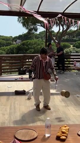 A young person with dark hair, wearing a pink and white striped shirt, white cargo pants, and brown boots, is in the center of a wooden deck, performing a yo-yo trick. They are holding a yo-yo with red strings and have a brown crossbody bag. To the left of the performer, a small white rabbit stands on its hind legs. In the background, three adults are standing and observing. The deck has a sloped, clear roof overhead, decorated with red and white striped bunting. Beyond the deck is a railing overlooking a lush, green landscape with trees and houses. A wooden table is in the foreground, with a bottle of water and a plate of pastries on it. The scene appears to be outdoors on a sunny day.